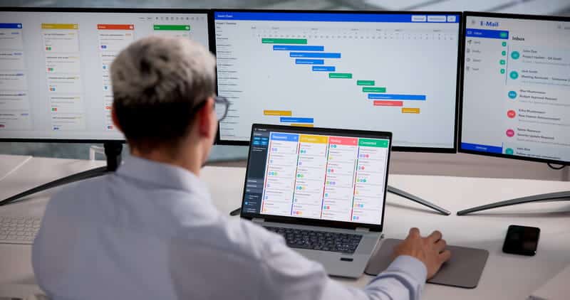 A person working on his laptop with three monitors withproject management tasks on screen.